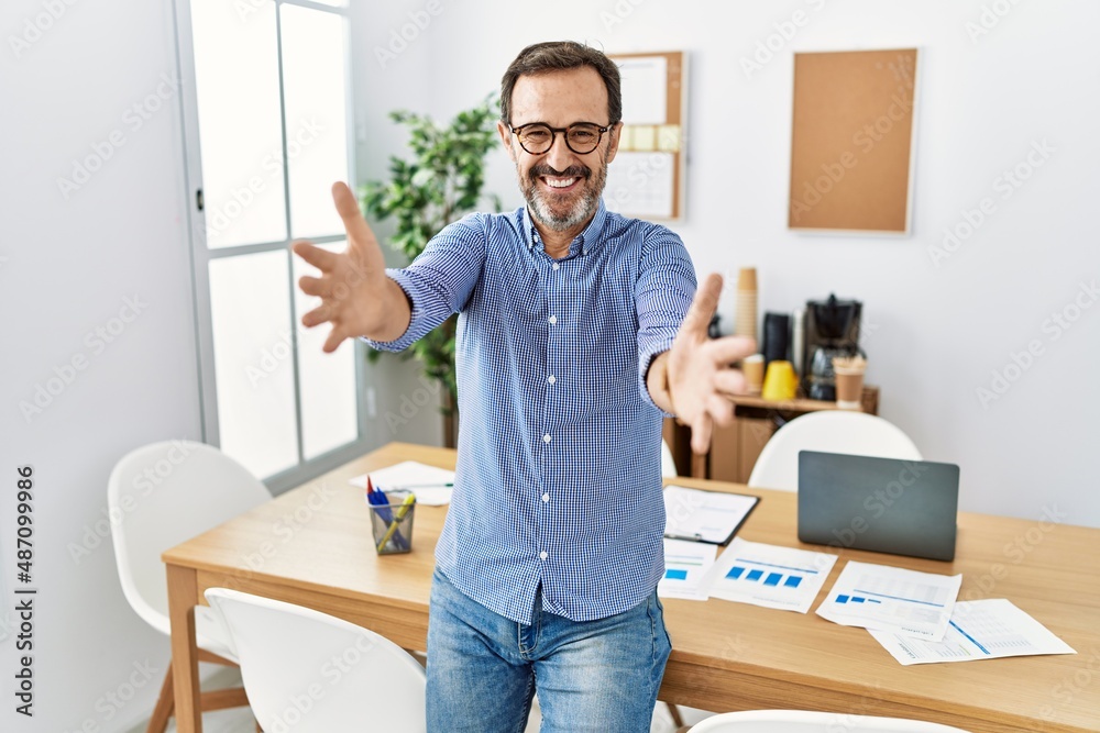 Middle age hispanic man with beard wearing business clothes at the office looking at the camera smiling with open arms for hug. cheerful expression embracing happiness. Middle age hispanic man with beard wearing business clothes at the office looking at the camera smiling with open arms for hug. cheerful expression embracing happiness.