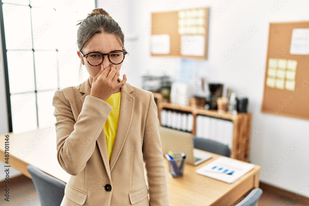 Young brunette teenager wearing business style at office smelling something stinky and disgusting, intolerable smell, holding breath with fingers on nose. bad smell - obrazy, fototapety, plakaty 