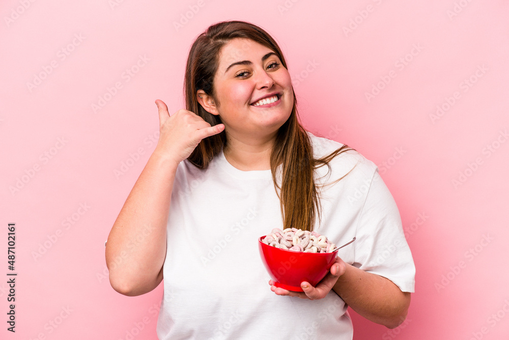 Young caucasian overweight woman holding a bowl of cereals isolated on pink background showing a mobile phone call gesture with fingers. Young caucasian overweight woman holding a bowl of cereals isolated on pink background showing a mobile phone call gesture with fingers.