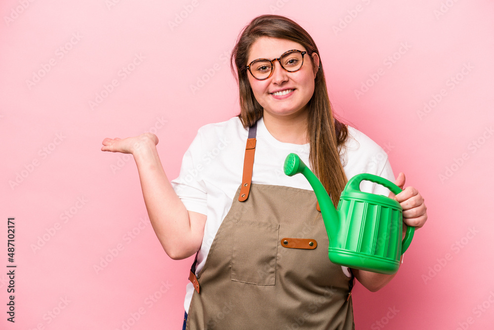 Young gardener caucasian overweight woman holding watering can isolated background showing a copy space on a palm and holding another hand on waist.