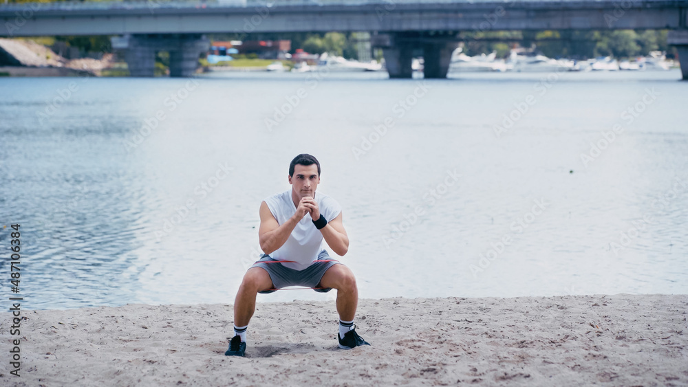 young athletic sportsman doing squat exercise on river shore