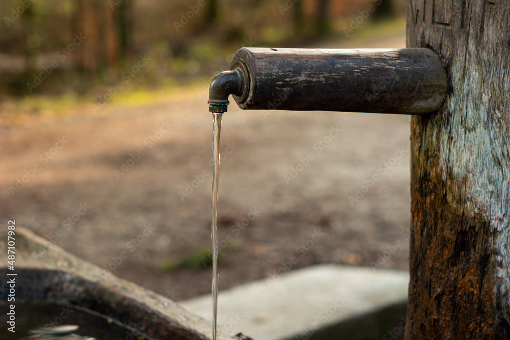 Old rustic drinking water fountain in the forest. Wooden construction, rusty metal pipe and faucet, streaming water. Close up shot, shallow depth of field, no people