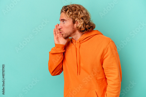 Young caucasian man isolated on blue background is saying a secret hot braking news and looking aside
