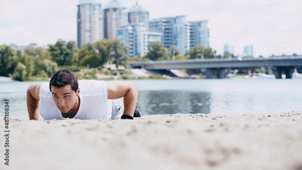 young sportsman doing push ups on sand near river in city