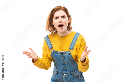 Shocked face red-haired girl looks surprised with open mouth spreads hands on white studio background