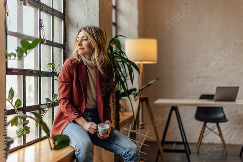 Thoughtful blonde woman, gazing outside the window, sitting and sipping tea.