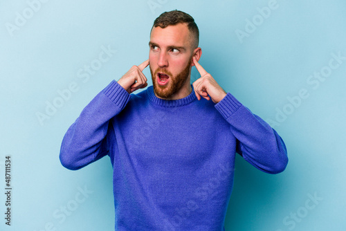 Young caucasian man isolated on blue background covering ears with fingers, stressed and desperate by a loudly ambient.