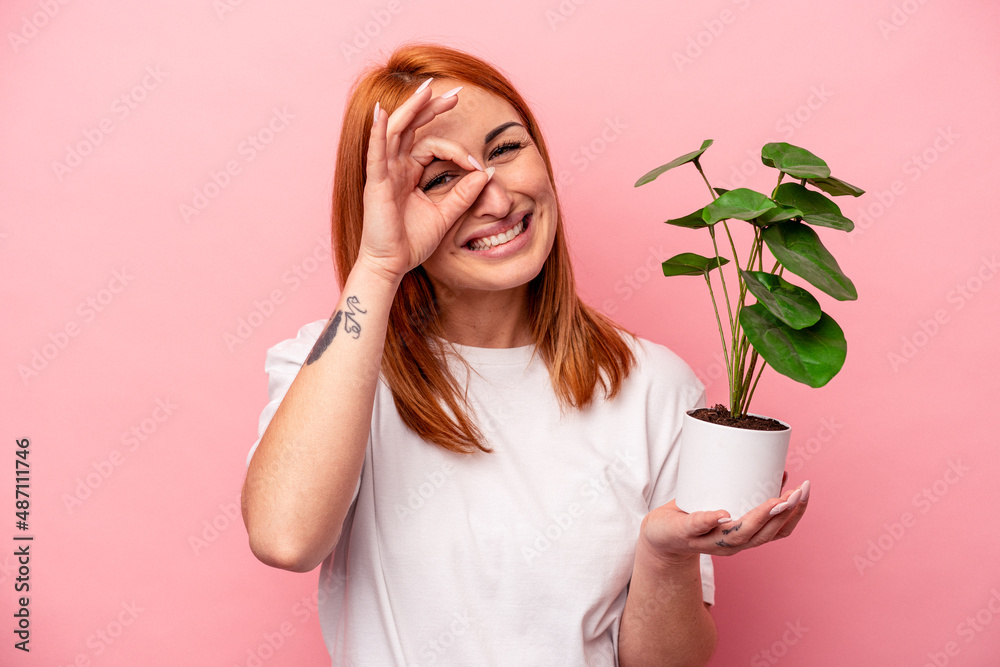 Young caucasian woman holding a plant isolated on pink background Young caucasian woman holding a plant isolated on pink background excited keeping ok gesture on eye.