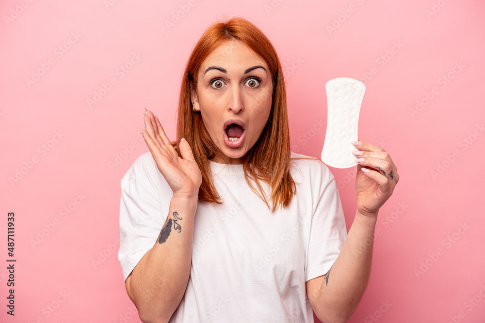 Young caucasian woman holding sanitary napkin isolated on pink background surprised and shocked. Young caucasian woman holding sanitary napkin isolated on pink background surprised and shocked.
