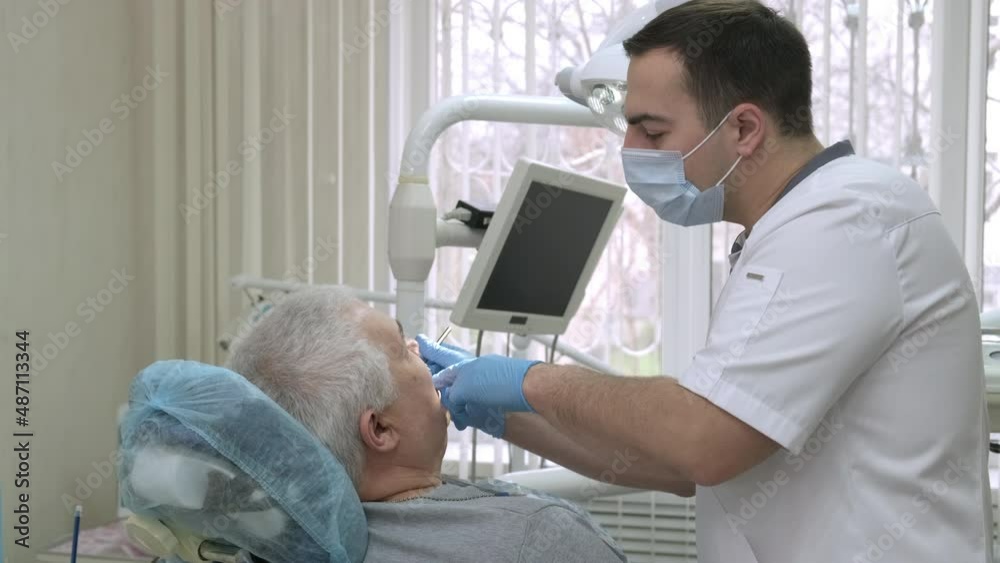 Senior man getting treatment at dental office. Young male dentist in protective mask working with elderly patient in clinic. Doctor working with client's teeth. Healthcare and medicine concept Senior man getting treatment at dental office. Young male dentist in protective mask working with elderly patient in clinic. Doctor working with client's teeth. Healthcare and medicine concept