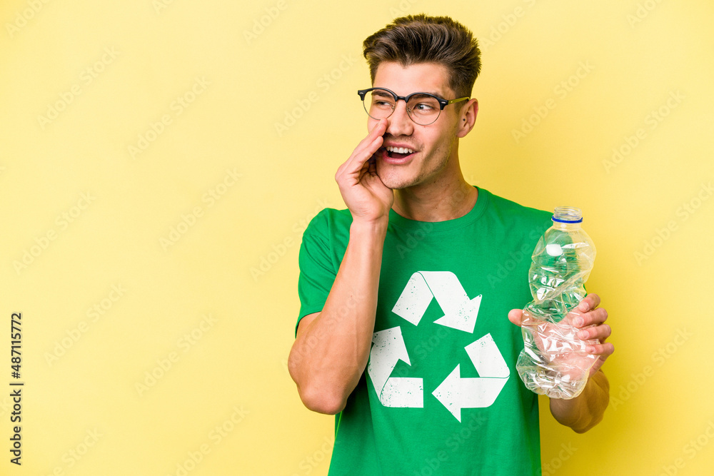 Young caucasian man holding a bottle of plastic to recycle isolated on yellow background is saying a secret hot braking news and looking aside