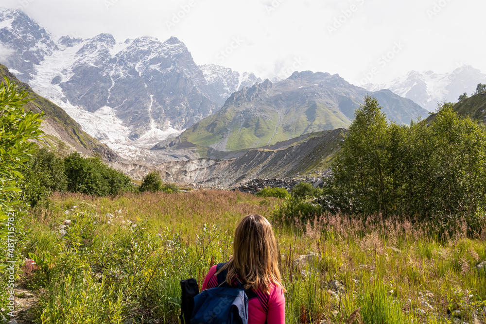 A woman hiking and enjoying the panoramic view on the Shkhara Glacier in the Greater Caucasus Mountain Range in Georgia, Svaneti Region, Ushguli. Wanderlust, Alpine pasture. Blooming flower.