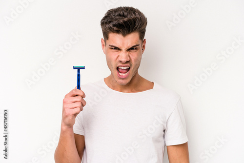 Young caucasian man shaving his beard isolated on white background screaming very angry and aggressive.