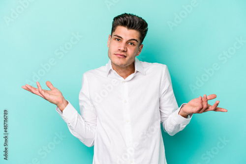 Young caucasian man isolated on blue background doubting and shrugging shoulders in questioning gesture.
