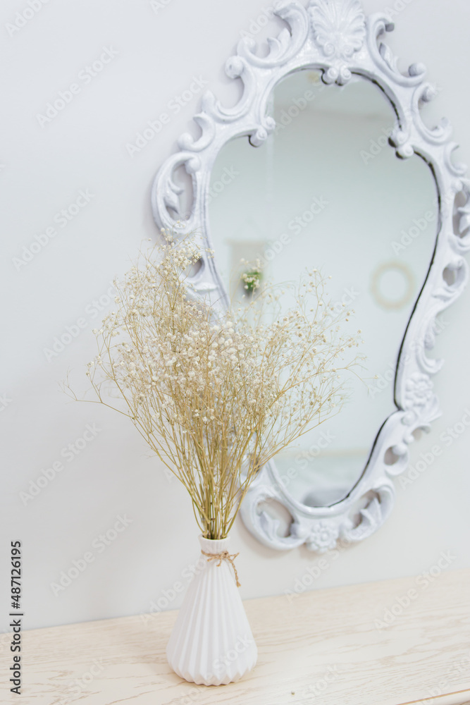 A bouquet of dry wildflowers stands in a white vase on a boudoir table by a vintage mirror.