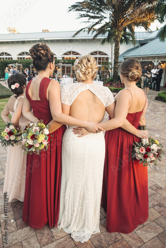 Bridesmaids and bride in a long white gown standing arm in arm holding bouquets of flowers with backs to the camera wearing long red dresses 