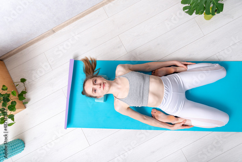 Relaxed woman lying on fitness mat and foam blocks. Girl doing fitness exercise at home. Supta Virasana. Top view photo