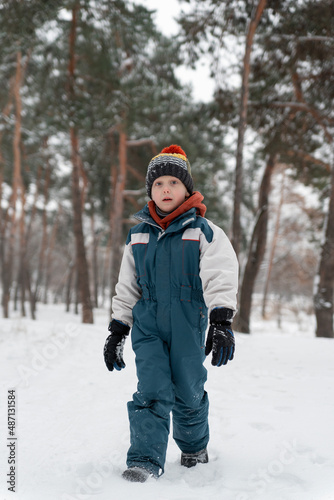 Funny child stands in snowy forest and looking at the camera. Boy stands against of beautiful winter landscape