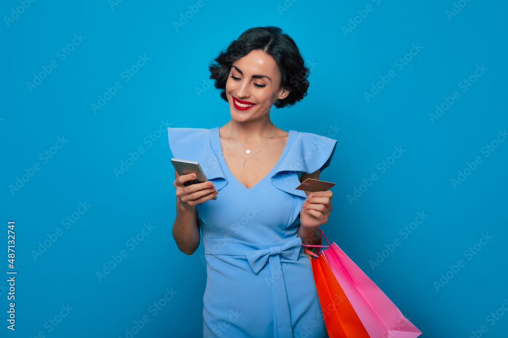 Online shopping. Close up portrait of smiling cute stylish brunette woman with shopping bags and credit card while she holds smart phone and makes purchases in app