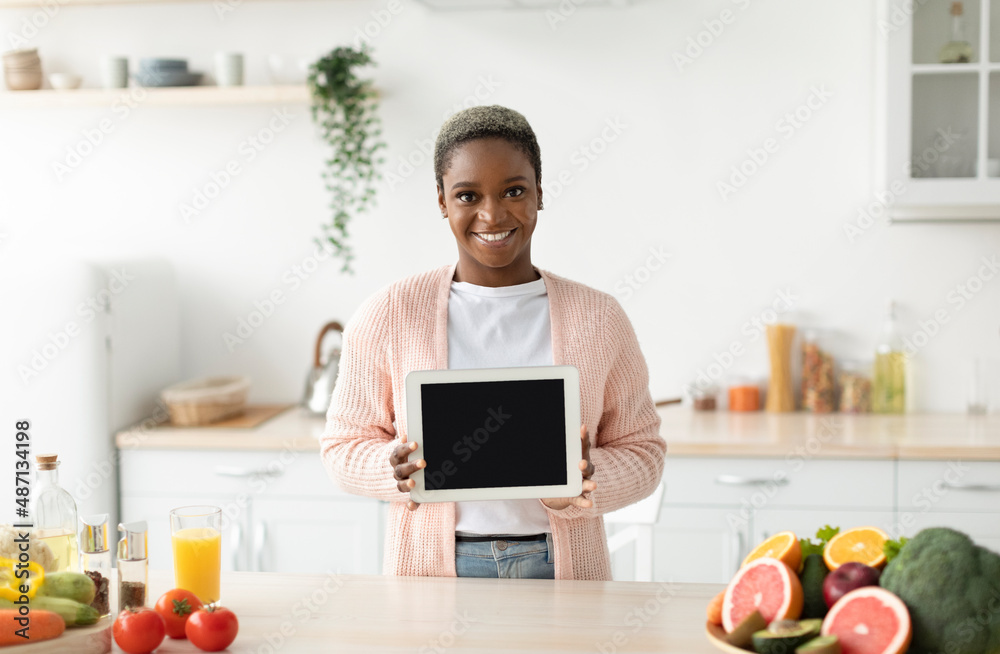 Smiling millennial african american woman in casual show tablet with blank screen in kitchen interior