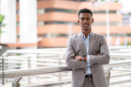 Young African businessman wearing suit standing in the city with building background. Crossed arm and smile. Business and lifestyle concept. Standing at walkway in the city. Portrait of worker