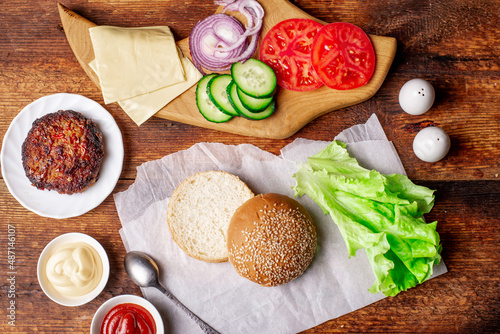 Cooking hamburger or cheeseburger. Different ingredients for a classic hamburger. Grilled meat, vegetables, greens, sauces near a sesame bun. Wooden background. Hamburger day.