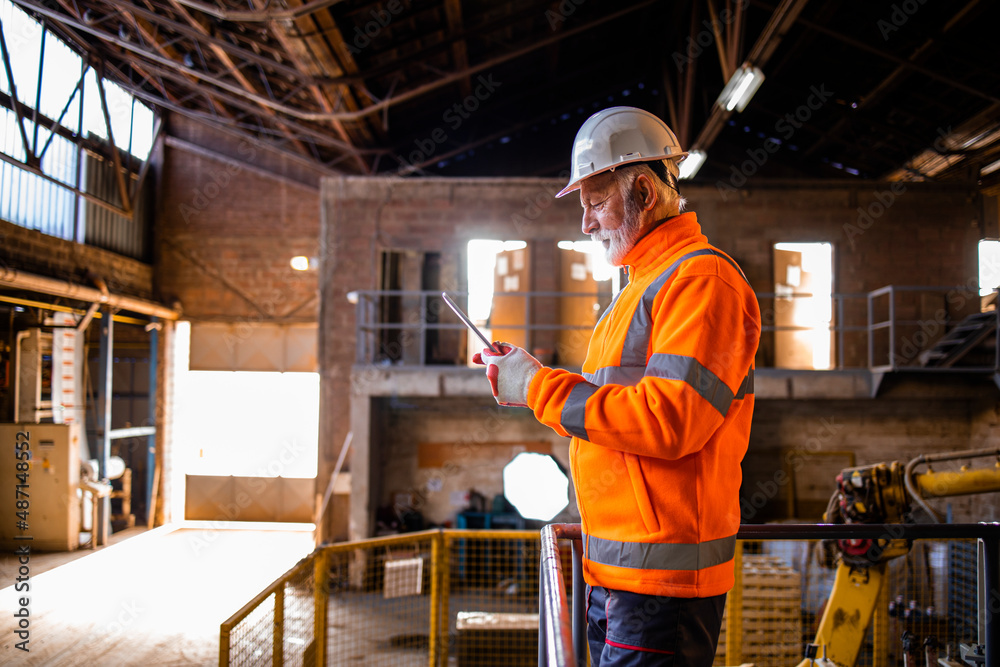Factory worker in safety uniform and white hard hat standing inside manufacturing factory and controlling production machines. Factory worker in safety uniform and white hard hat standing inside manufacturing factory and controlling production machines.