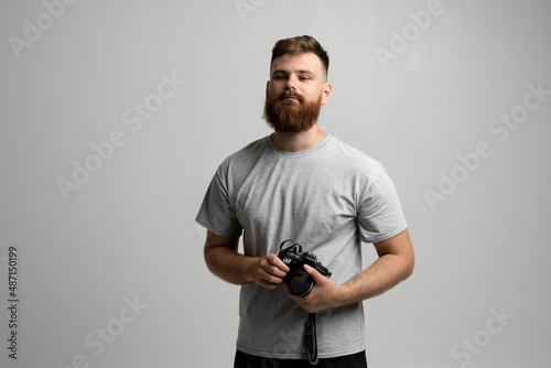 Portrait of bearded professional photographer in a grey t-shirt with dslr camera looks straight into the camera isolated on gray background.