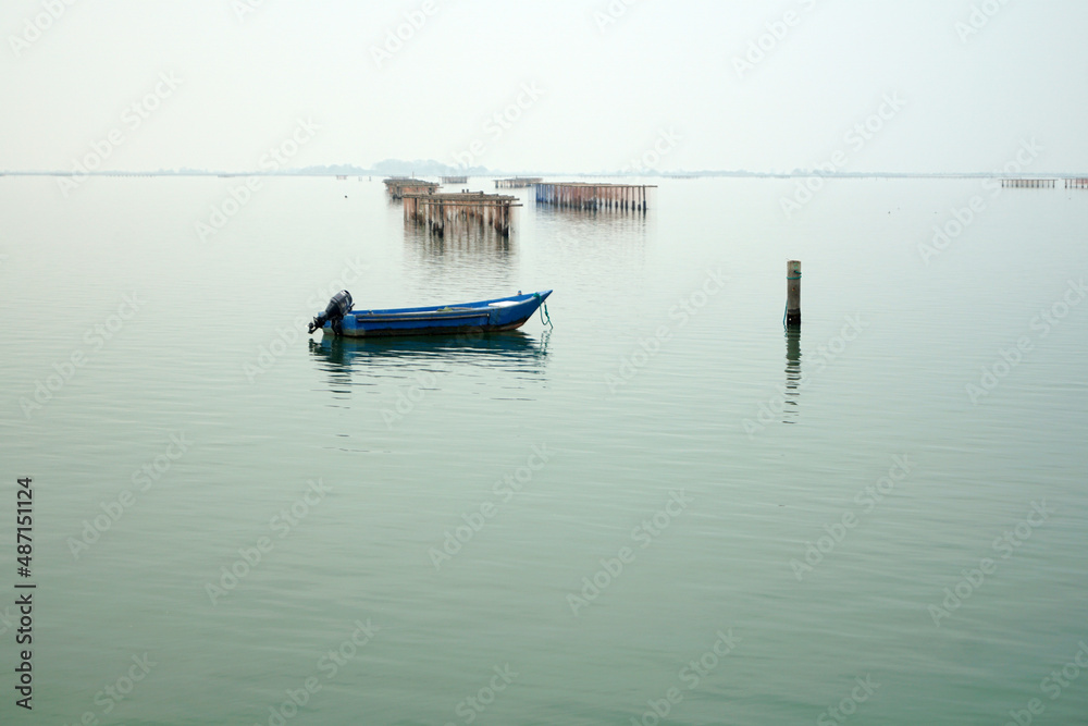 Barca azzurra immersa in atmosfera calma e misteriosa del mare piatto con sfondo allevamenti di vongole e cozze nel mare adriatico presso Chioggia Venezia Italia