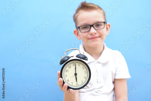 a boy on a blue background with glasses holds an alarm clock in his hands. it is difficult for a child to wake up in the morning, he is tired. you need to drink vitamins for vivacity and immunity