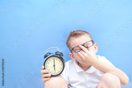 a boy on a blue background with glasses holds an alarm clock in his hands. it is difficult for a child to wake up in the morning