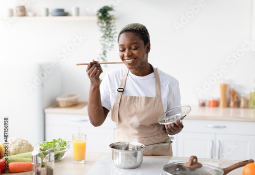 Smiling millennial african american woman in apron cooking at home and tasting dish in kitchen interior