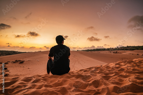 Young asian man wearing hat sitting on sand dune during the sunset
