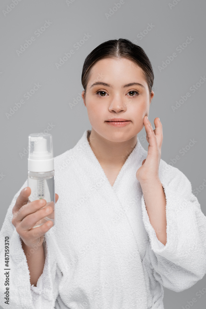 Young woman with down syndrome in bathrobe holding cleaning foam and touching face isolated on grey Young woman with down syndrome in bathrobe holding cleaning foam and touching face isolated on grey
