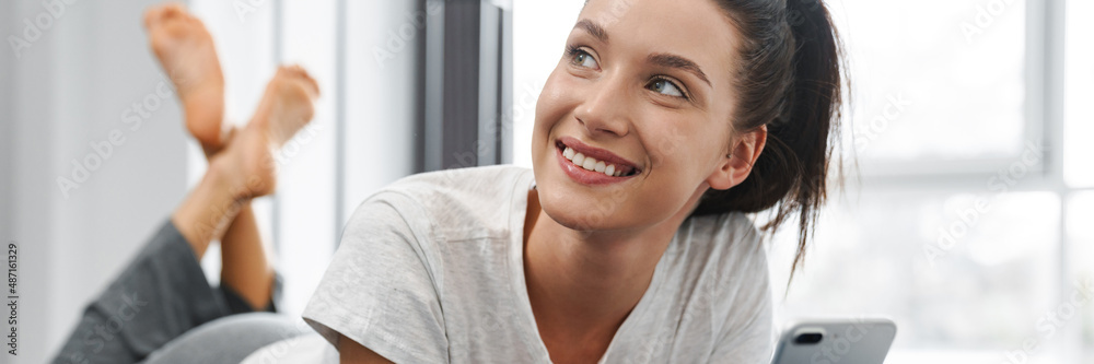 European smiling woman using mobile phone while lying on bed at home European smiling woman using mobile phone while lying on bed at home