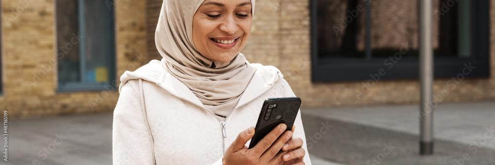 Middle eastern woman in hijab using mobile phone and smiling outdoors Middle eastern woman in hijab using mobile phone and smiling outdoors