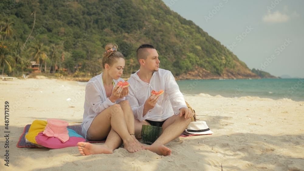 The cheerful love couple holding and eating slices of watermelon on tropical sand beach sea. Romantic lovers two people caucasian spend summer weekend in outdoor. Hat, backpack white shirt beachwear.
