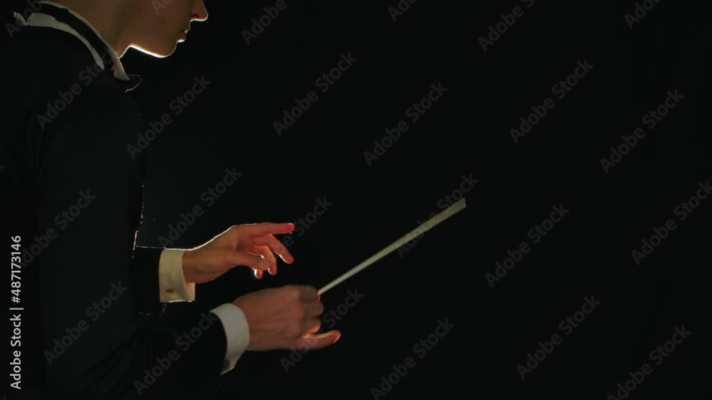 Symphony orchestra conductor wearing suit is directing musicians with movement of baton on black background with lights. Conducting, directing a musical performance with visible gestures. Close up.