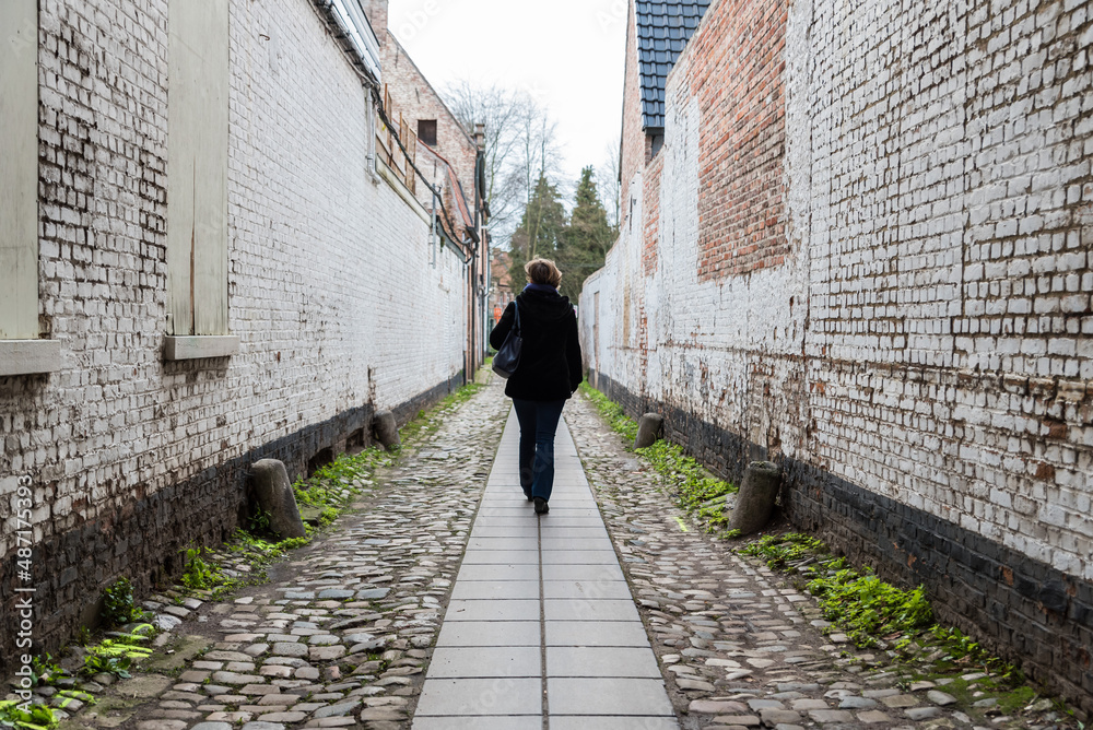 Woman walking through a small alley with a paved road an old bricked walls at the beguinage of Dendermonde, Flanders, Belgium Woman walking through a small alley with a paved road an old bricked walls at the beguinage of Dendermonde, Flanders, Belgium