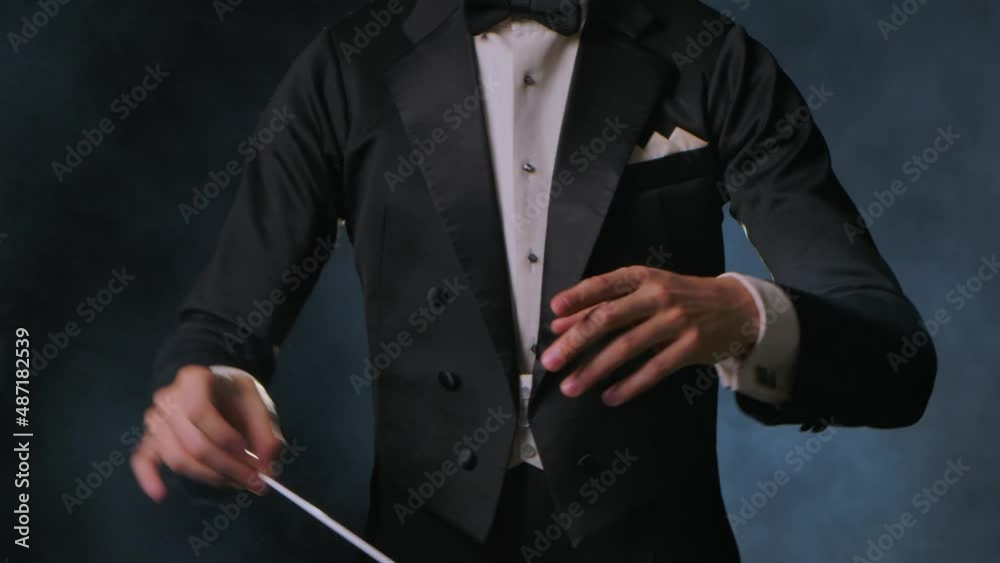 Concert of classical music under direction of an orchestra conductor. Man waves his baton to synchronize and conducting musicians in theater. Black background with smoke and backlight. Close up.