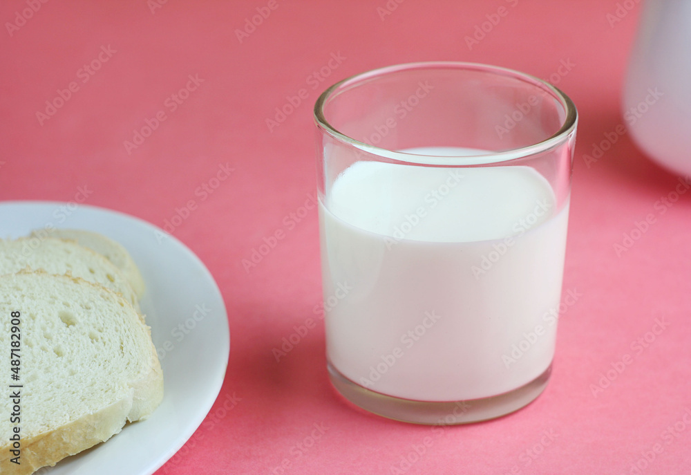 A glass of milk stands on a pink table, next to it lies bread in a white plate. Behind is a milkman A glass of milk stands on a pink table, next to it lies bread in a white plate. Behind is a milkman