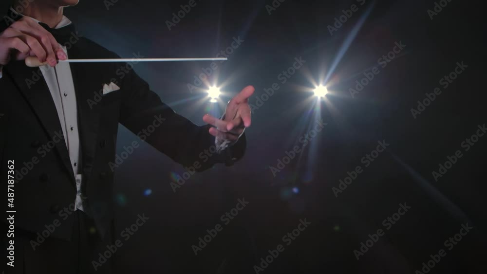Moving hands of an orchestra conductor directing musicians with movement of baton on black background with lights. Conducting, directing classical musical performance with visible gestures. Close up. Moving hands of an orchestra conductor directing musicians with movement of baton on black background with lights. Conducting, directing classical musical performance with visible gestures. Close up.