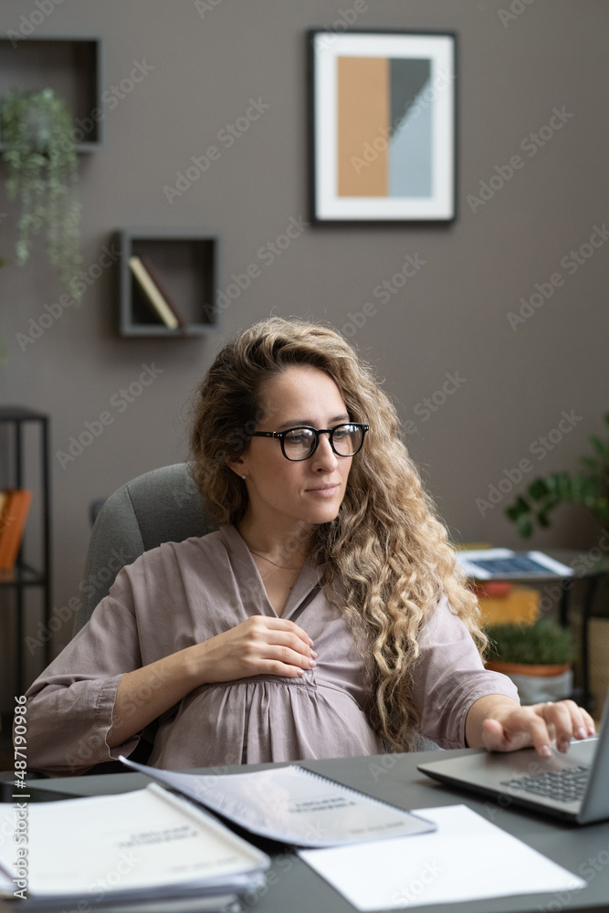 Young pregnant woman sitting at her workplace and typing on laptop during her work at office Young pregnant woman sitting at her workplace and typing on laptop during her work at office