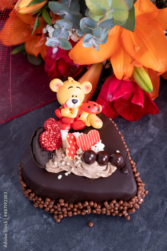 Heart-shaped chololate cake accompanied by a teddy bear and a bouquet of flowers, on a slate background