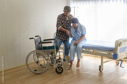 Senior man helping cancer patient woman wearing head scarf moving to wheelchairs at hospital, health care and medical concept