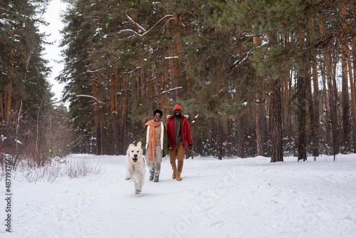 Happy African couple in warm clothing walking along the path in winter forest with their running dog