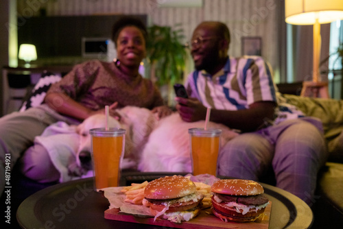 Close-up of burgers and soda on the table with African couple watching tv together on the sofa in the background