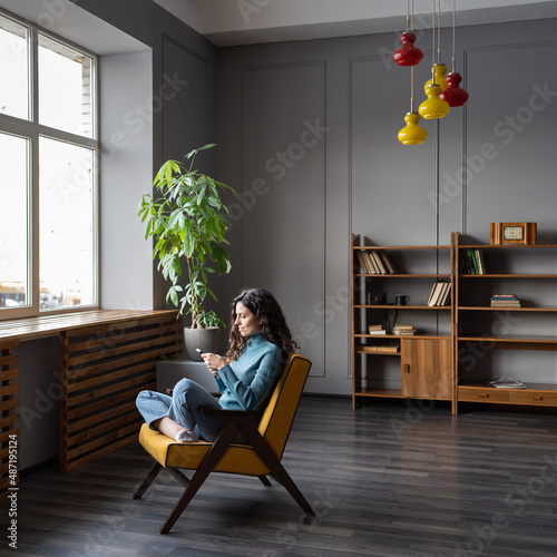 Happy young woman sit relaxed in comfortable armchair at home hold smartphone checking email or social media messenger. Smiling millennial female rest in chair browse web internet on modern cellphone photo