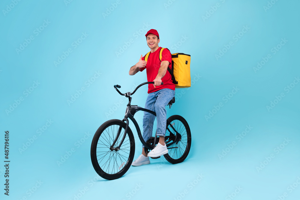 Fototapeta premium Cheerful Male Courier With Bike Gesturing Thumbs Up, Blue Background