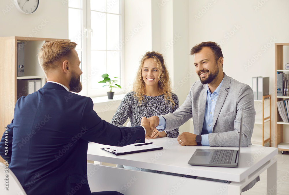 Happy young couple shaking hands with real estate agent after signing contract at his office. Clients smiling and exchanging handshake with realtor. Happy family thanking loan advisor for consultation - obrazy, fototapety, plakaty 